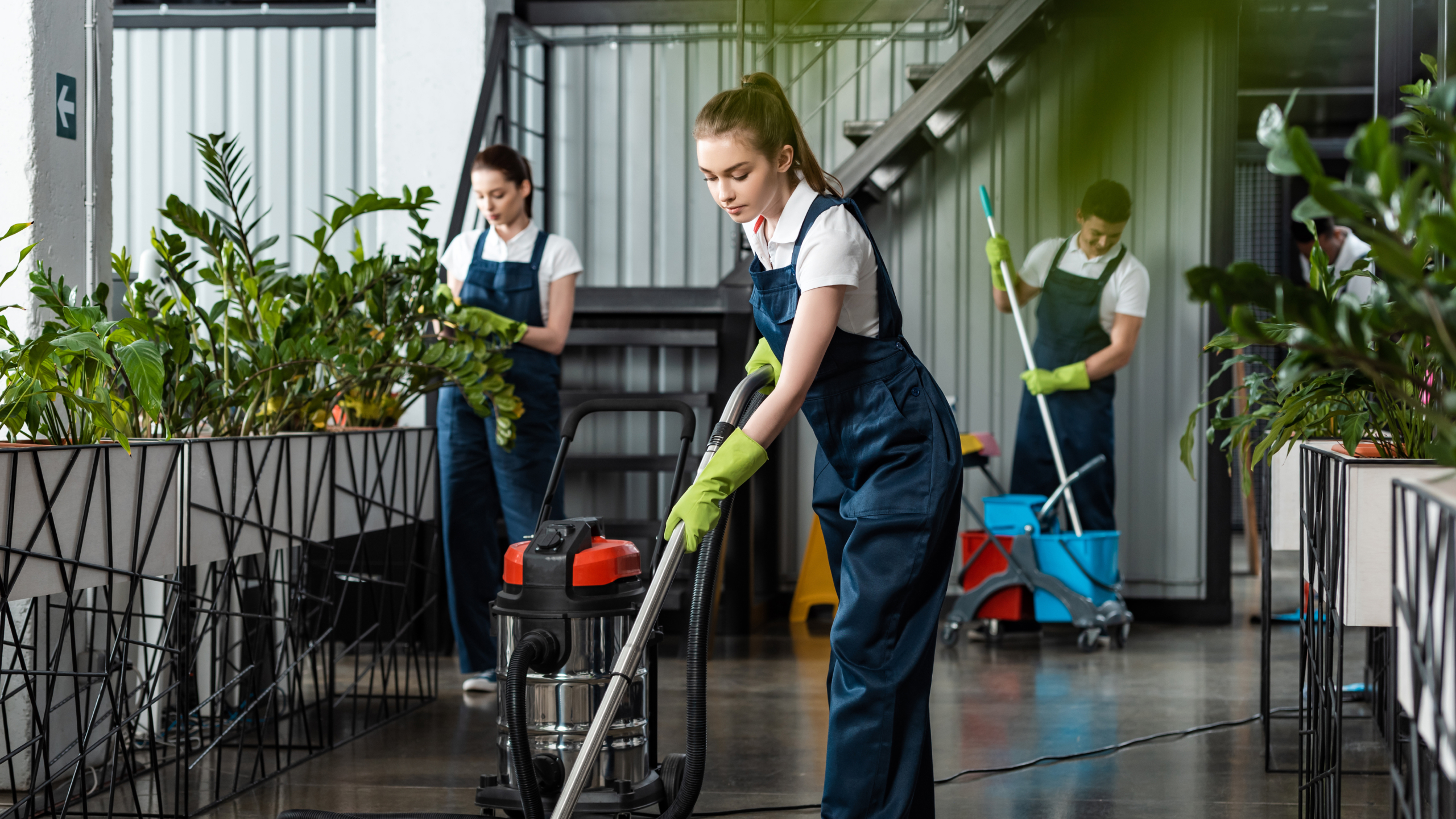 Three individuals in uniforms are cleaning a modern indoor space with plants. The foreground shows a woman using a vacuum cleaner, while two others are cleaning nearby with mops and equipment. Everyone is wearing gloves and focused on their tasks.